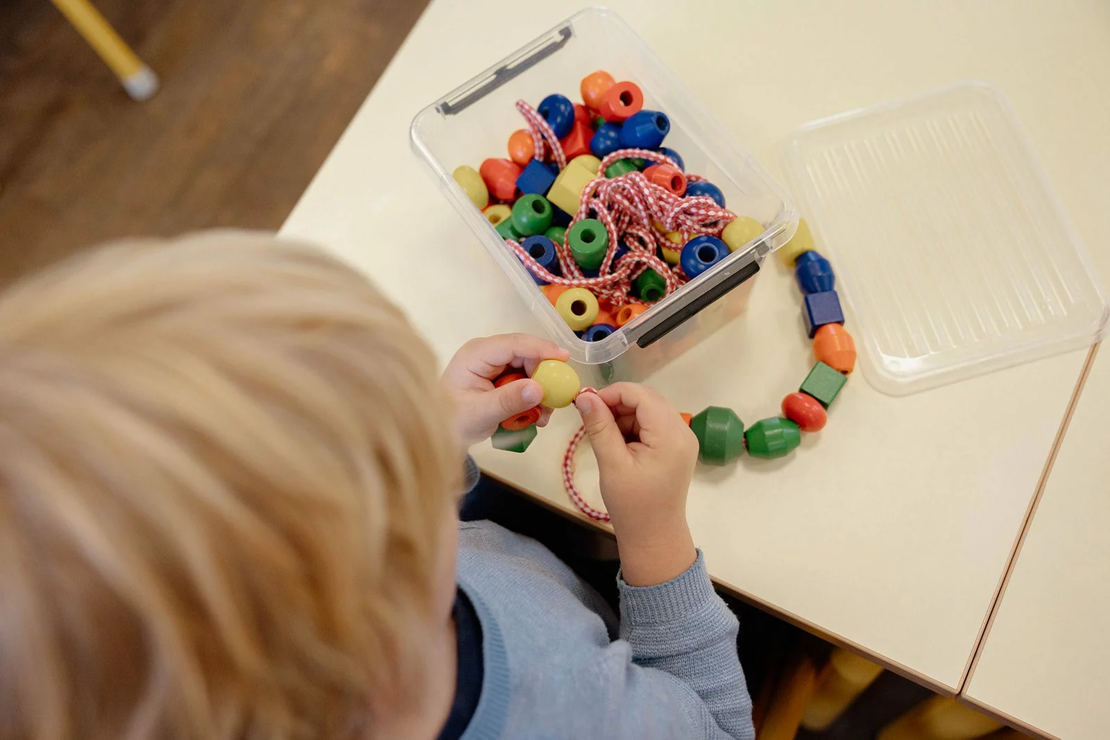 Un enfant de maternelle, vu de dos, enfile des perles de bois sur un lacet