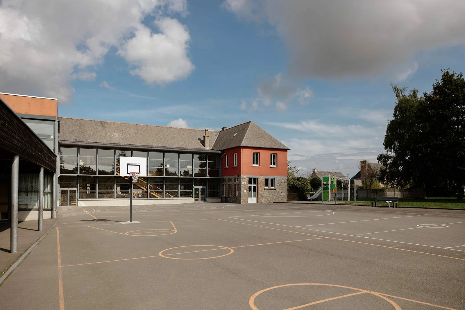 La cour de l’école vide sous un ciel bleu : le panier de basket, la façade vitrée et l’aire de jeux
