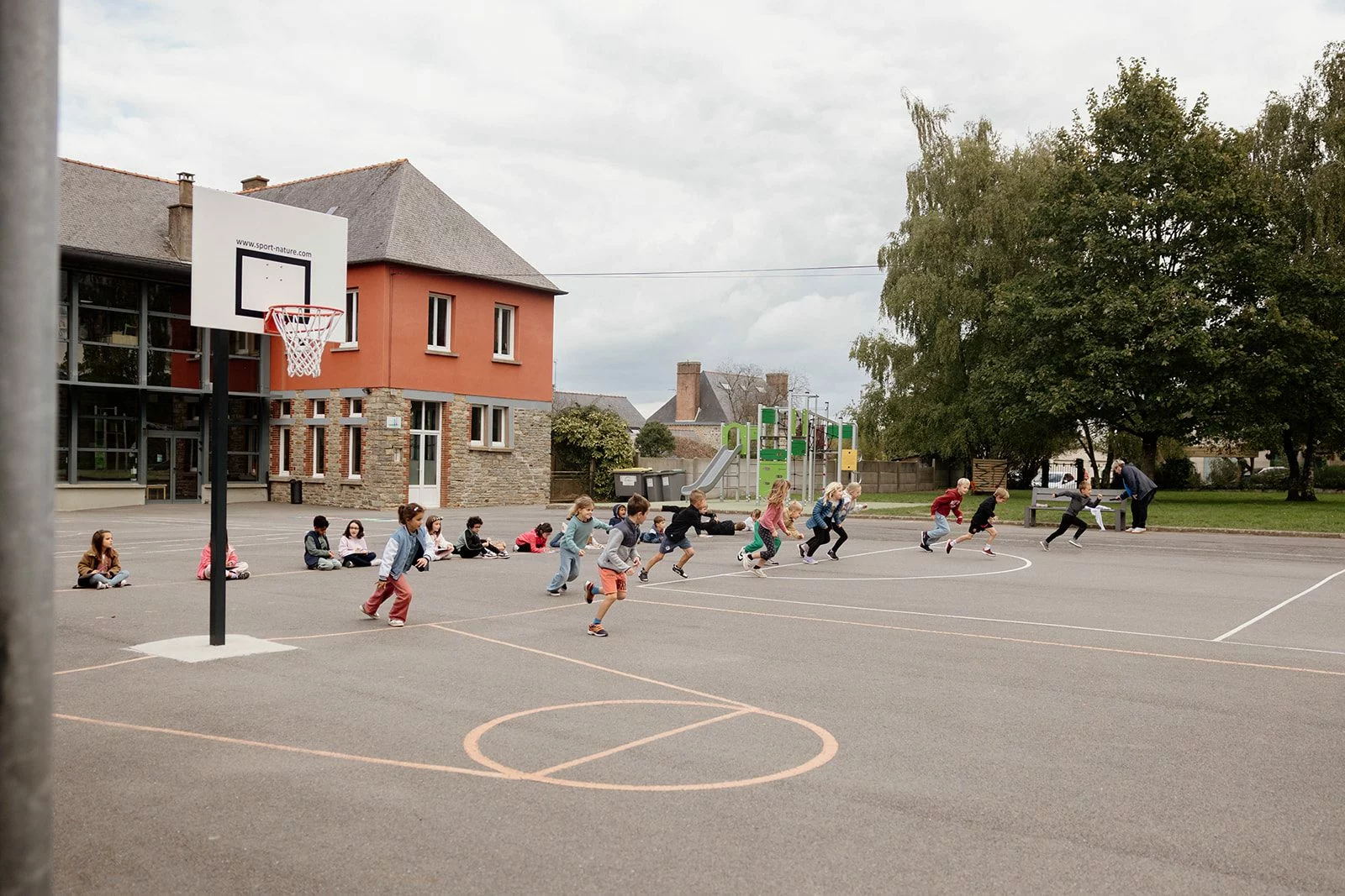 Des élèves s'élancent pour une course dans la cour de l'école, sous le regard d'une adulte