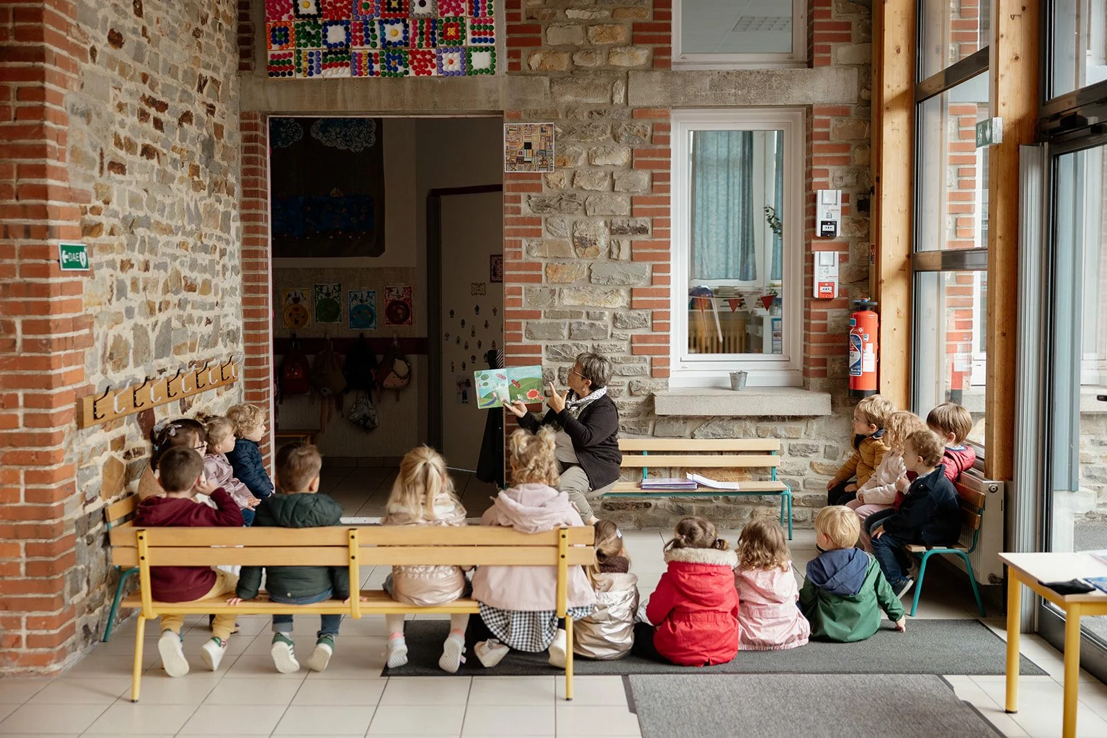 Une adulte lit un album illustré à un groupe d'enfants assis dans le hall de l'école