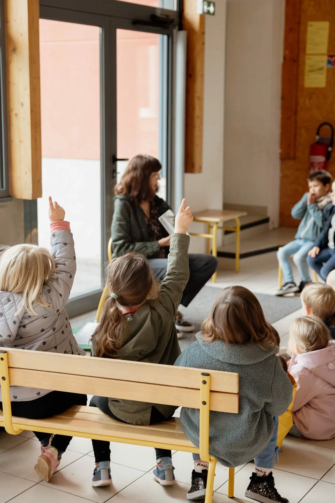 Des enfants assis en cercle lèvent la main pendant qu'une adulte leur lit un album