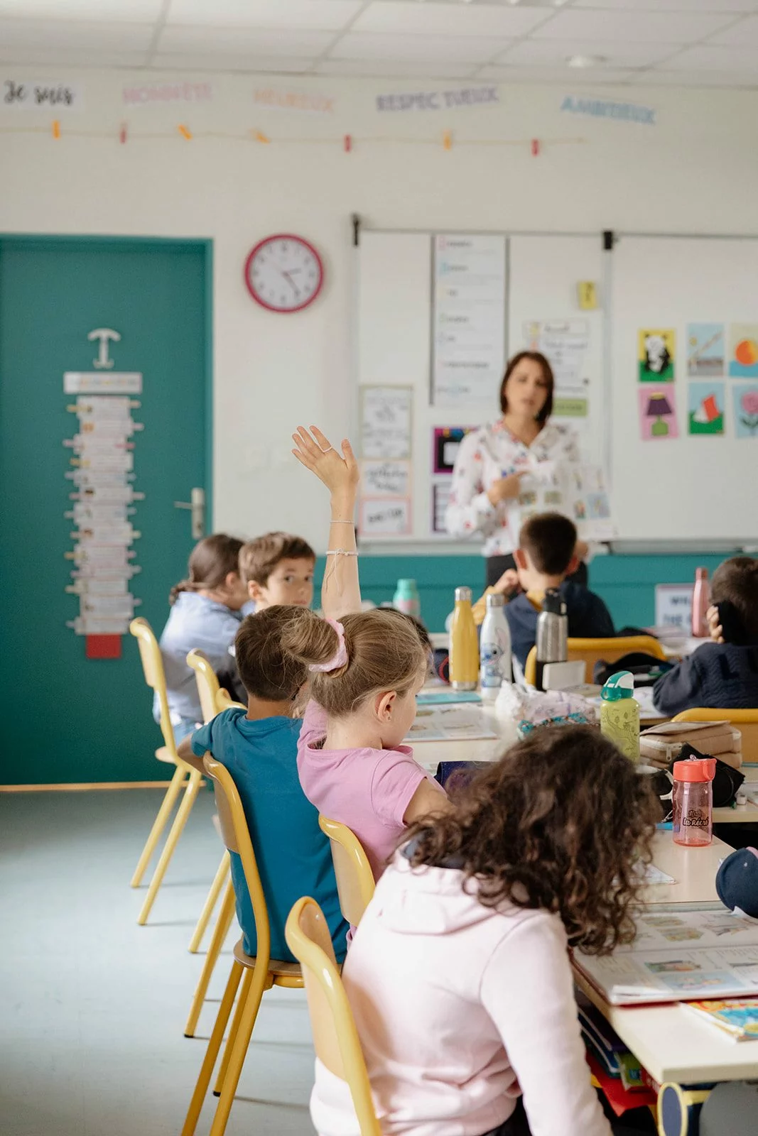 Une élève lève la main dans une classe d'élémentaire, l'enseignante devant le tableau