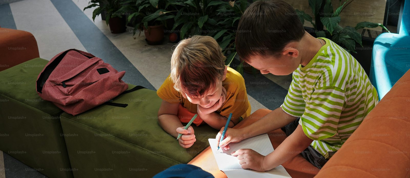 Deux enfants assis cote a cote ecrivent sur une feuille, leurs cartables poses pres d eux
